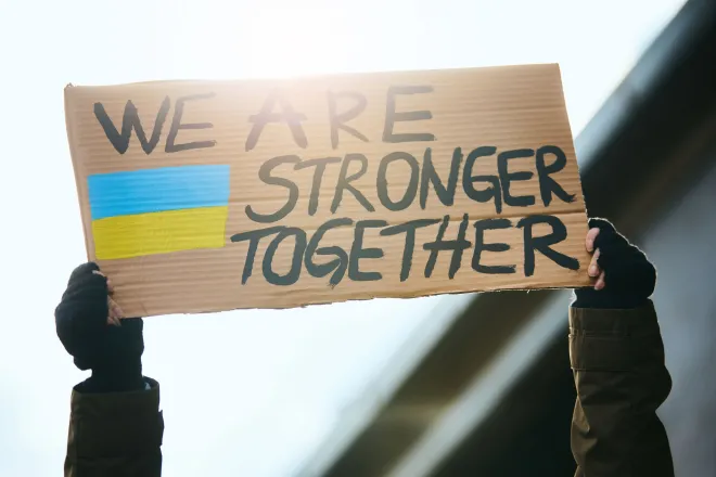 Foto van twee handen die een protestbord in de lucht houden met erop geschreven: We are stronger together. Aan de zijkant een geschilderde blauw-gele vlag. 